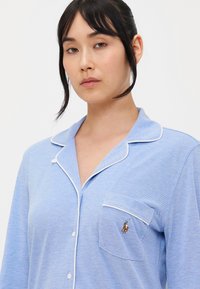 Haut de pyjama bleu clair avec une finition texturée, des passepoils blancs le long du col et de la poche, et un petit détail brodé sur la poche poitrine.