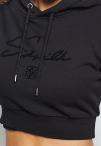 Sudadera corta negra con cordones y un sutil bordado del logo negro de SikSilk en el pecho, llevada en la parte superior del torso de una persona.