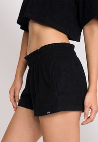 Pantaloni shorts neri in spugna con vita arricciata elastica, tasche laterali e un piccolo etichetta con logo sull'orlo. Texture morbida e design semplice.