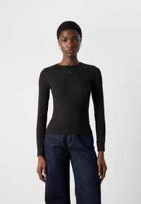Black long-sleeve ribbed top with a round neckline. Features a small logo detail on the chest. Worn with dark blue wide-leg jeans.