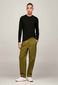 Svart langermet skjorte, olivengrønne plisserte bukser og hvite joggesko med teksturerte såler. Modell står med en hånd i lommen.