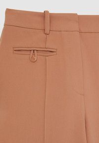 Pantalones de color durazno con una textura suave, que cuentan con un lazo para cinturón, un detalle de botón en el bolsillo trasero y costuras visibles.