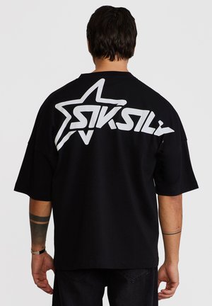 Mann mit kurzen dunklen Haaren trägt ein schwarzes übergroßes T-Shirt mit einem großen weißen Stern und stilisiertem "SikSilk"-Schriftzug auf dem Rücken.