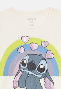 Kremowa koszulka z nadrukiem kreskówkowej postaci o niebiesko-szarym ciele, otoczonej trzema brokatowymi różowymi sercami i tęczowym tłem.