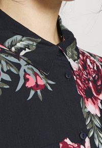 Camicia a bottoni floreale in tessuto nero con motivi di fiori rosa e verdi, colletto e dettagli intrecciati lungo il pattino dei bottoni.