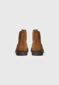 Bottines en cuir suédé marron avec des languettes, semelles épaisses noires, vues de l'arrière sur un fond blanc uni.