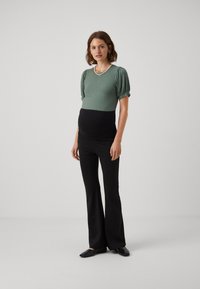Haut court à rayures vertes avec un design ajusté, associé à un pantalon de maternité noir évasé. Des chaussures noires à enfiler complètent la tenue.