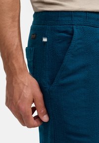 Türkisfarbene, strukturierte Hose mit elastischem Bund, Fronttaschen und kontrastierenden Stoffflicken. Eine Nahaufnahme einer Hand, die den Stoff berührt.