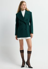 Abrigo de mezcla de lana verde, de doble botonadura y con grandes solapas, con cuatro botones grandes, combinado con un vestido mini blanco y medias altas negras.