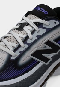 Primo piano di una scarpa da corsa grigia e nera con lacci bianchi, dettagli viola e un grande logo "N" sul lato.