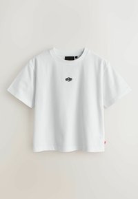 Camiseta blanca de algodón de manga corta con un pequeño logo negro bordado en el pecho y una etiqueta roja en la costura lateral izquierda.