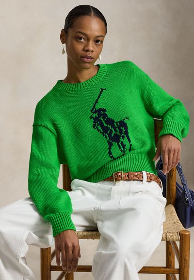 Πράσινο πλεκτό πουλόβερ με το λογότυπο ενός παίκτη πόλο σε σκούρο μπλε χρώμα, συνδυασμένο με λευκό παντελόνι και πλεκτή καφέ ζώνη. Ουδέτερο φόντο.