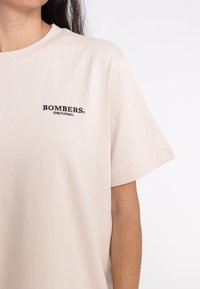 Beige Baumwoll-T-Shirt mit kurzen Ärmeln und Rundhalsausschnitt, mit schwarzem besticktem Text "BOMBERS ORIGINAL" auf der linken Brust.
