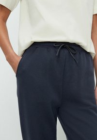 Marina joggerbyxor med dragsko i midjan, sidfickor och mjukt tyg, tillsammans med en ljusbeige kortärmad skjorta.