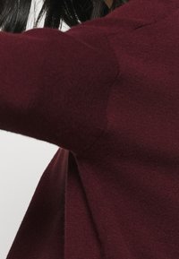 Maglione a maglia borgogna che mostra un tessuto texturizzato, taglio triangolare sul lato e un primo piano della cucitura della manica. Design morbido e informale.