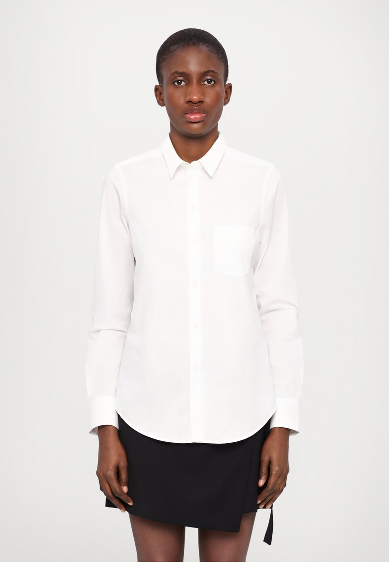 Camicia bianca con bottoni, maniche lunghe, orlo arrotondato e una piccola tasca sul petto, abbinata a una gonna nera. Tessuto liscio e design rigoroso.