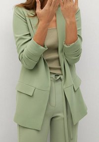 Blazer vert clair avec une coupe ajustée, deux poches avant et une texture douce, associé à un débardeur beige à côtes et un pantalon assorti.