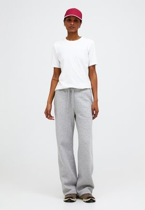 Femme portant une casquette rouge, un t-shirt blanc à manches courtes, un pantalon de survêtement gris ample et des baskets marron, debout devant un fond uni.