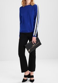 Pull bleu avec des rayures blanches sur les manches, pantalon noir, pochette noire avec une fermeture éclair dorée, et chaussures à talons hauts noires.