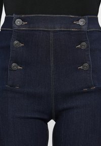 Jeans en denim bleu foncé taille haute avec une texture lisse, dotés de quatre boutons noirs plats et de coutures beiges visibles sur le devant.