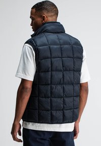 Marineblauer Steppweste, mit durchgestepptem Design, hohem Kragen, ärmellos, aus leichtem Material. Getragen über einem hellgrauen Shirt mit quadratischen Mustern.