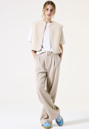 Un gilet en polaire beige sur un t-shirt blanc, associé à un pantalon beige clair et des chaussures de sport bleues avec des accents jaunes.
