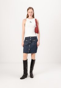 Bela brezrokavnica s logotipom, temno denim krilo na gumb spredaj in sijoči črni škornji do kolen. Rdeča torba s trakovi nošena na rami.