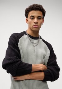 Rippstrickpullover mit kontrastierenden schwarzen und grauen Materialien, übergroßen Ärmeln und geschichteten silbernen Halsketten.