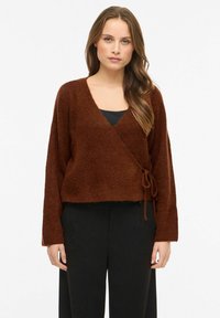 Brązowy, miękki sweter o kroju kopertowym z długimi rękawami, dekoltem w serek i regulowanym troczkiem w talii. Nosi się go na czarnym topie, w połączeniu z czarnymi spodniami.