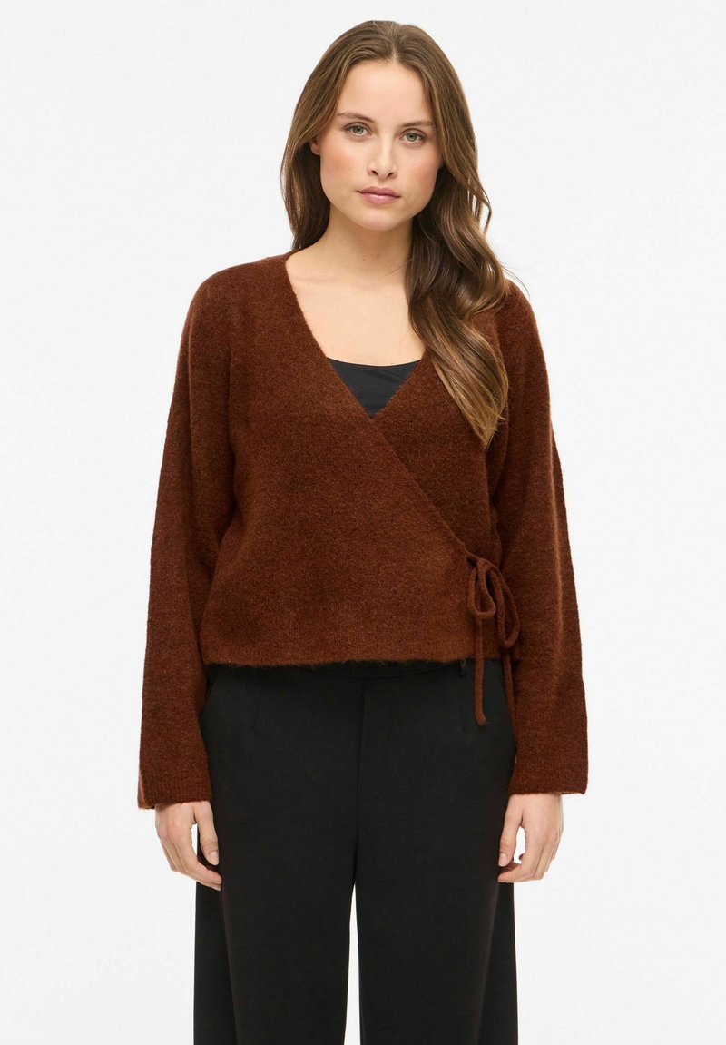 Brązowy, miękki sweter o kroju kopertowym z długimi rękawami, dekoltem w serek i regulowanym troczkiem w talii. Nosi się go na czarnym topie, w połączeniu z czarnymi spodniami.