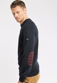 Mann trägt einen marineblauen Pullover mit Knöpfen an der Schulter und roten gepunkteten Ellbogenpatches, kombiniert mit beigen Hosen, und blickt nach unten.