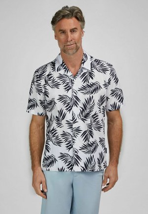 Homme portant une chemise blanche à manches courtes avec un imprimé feuilles de palmier noires et un pantalon bleu clair, debout devant un fond uni.