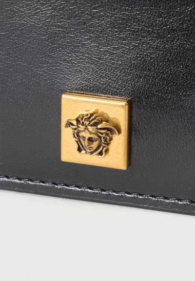 Surface en cuir noir avec couture visible et petit emblème carré en or représentant une tête de Méduse en relief.