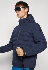 Ναυτικό μπουφάν με φερμουάρ, αποσπώμενη κουκούλα και ελαστικούς καρπούς; το μοντέλο κρατάει ένα μπαστούνι πεζοπορίας και φοράει σκούρα γυαλιά ηλίου.