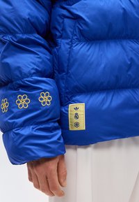 Χέρι που φοράει μπλε μπουφάν με κίτρινα κεντημένα λουλούδια στο μανίκι και κίτρινο ετικέτα με λογότυπα κοντά στη μέση σε λευκό φόντο.