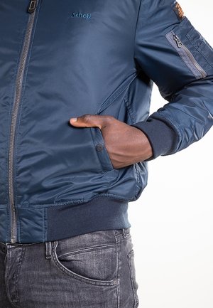 Gros plan d'une personne portant une veste bomber en nylon bleue Schott avec une main dans la poche latérale et un jean en denim gris.