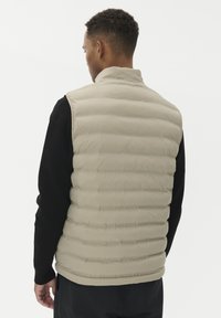 Mees seljaga vaatamas, kandes beeži tikitud vestit musta pika varrukaga särk ja tume püksid valge taustal.