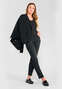 Svart blazer med texturerad yta, bärs över en svart topp, ihop med mörka jeans och glossiga svarta loafers. Stående pose mot en neutral bakgrund.