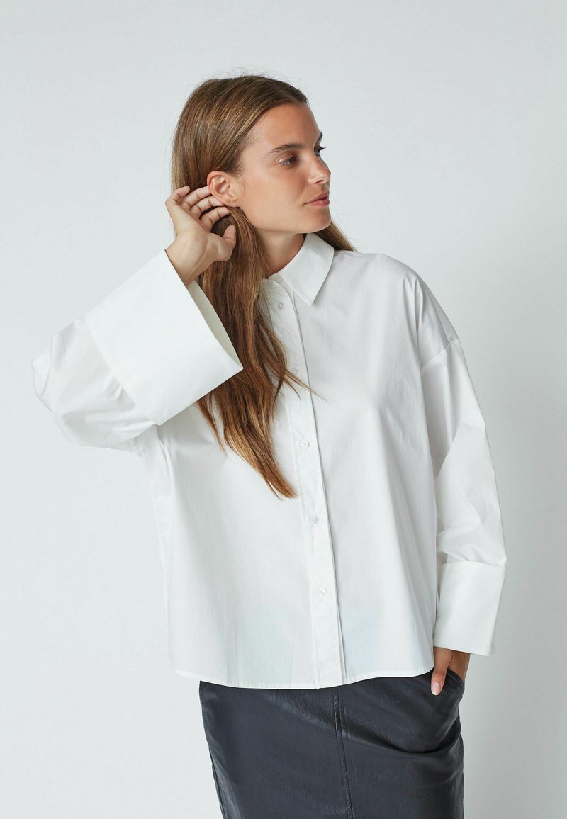Chemise blanche oversize à boutons, col pointu, manches larges et texture lisse, assortie à une jupe noire.