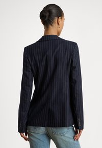 Moteris su kuodu šviesiais plaukais, vilkinti tamsiai mėlyną dryžuotą švarką ir mėlynas džinsus, stovinti nugara į žiūrovą ant vienspalvio balto fono.