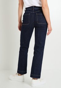 Jeans de denim azul oscuro de pierna recta con dos bolsillos traseros y costuras contrastantes. Combinados con zapatillas blancas, mostrados desde la parte de atrás.