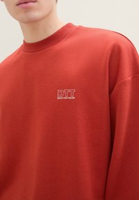 Felpa rossa in cotone con scollo a girocollo, caratterizzata da un piccolo logo bianco sul petto. Texture liscia e vestibilità rilassata con spalle scese.