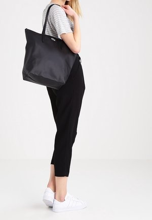 Femme portant un t-shirt à rayures, un pantalon noir court, des baskets blanches, tenant un grand sac cabas noir à l'épaule, debout devant un fond uni.