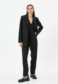 Complete abito nero su misura con blazer e gilet caratterizzati da scollatura a V e tre bottoni, abbinato a pantaloni larghi neri e mocassini.