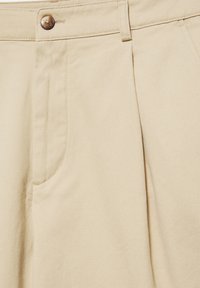 Pantalon beige taille haute avec fermeture par bouton, poche latérale et coutures visibles en gros plan.