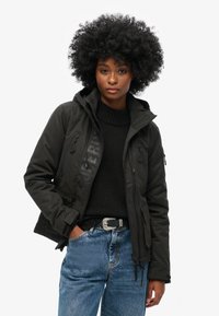 Schwarze Kapuzenjacke mit glänzendem Logo, Reißverschluss-Taschen und verstellbarem Saum. Getragen über einem schwarzen Pullover und kombiniert mit hellblauen Jeans.