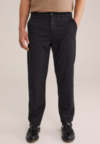Uomo che indossa pantaloni neri a taglio dritto, mocassini neri con nappine e una camicia beige, in piedi con una mano in tasca.