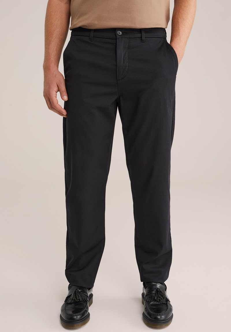 Uomo che indossa pantaloni neri a taglio dritto, mocassini neri con nappine e una camicia beige, in piedi con una mano in tasca.