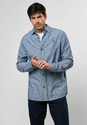 Camisa azul de manga larga con un patrón texturizado y un cuello clásico, llevada sobre una camiseta blanca, combinada con jeans oscuros.