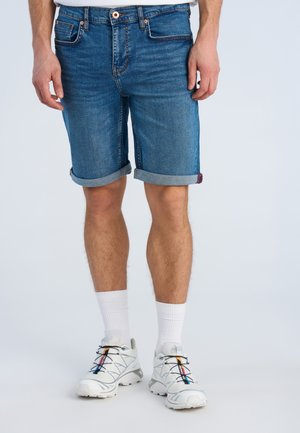 Mężczyzna w niebieskich jeansowych szortach z mankietami, białych sportowych skarpetach i białych butach do biegania z wielokolorowymi detalami, stoi na jednolitym tle.